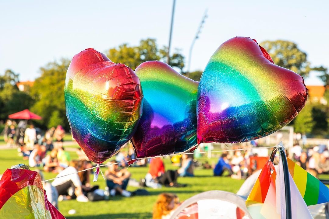 Drie glimmende regenboog ballonnen aan een touwtje. Op de achtergrond is een wei met mensen erop.