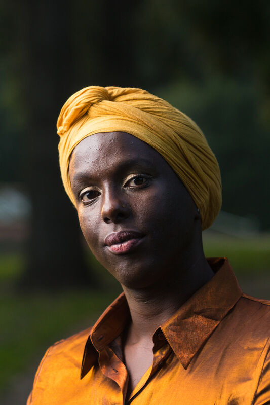 Portretfoto van spreker Olave Basabose - Olave Nduwanje, een zwarte vrouw met een okergele tulband om. Ze staat buiten bij een boom.