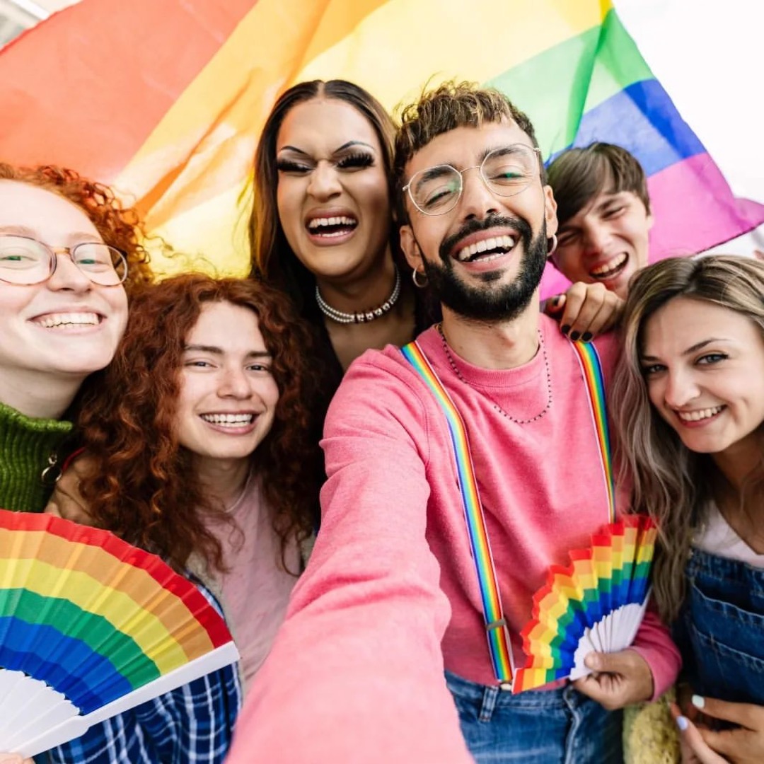 Kleurenfoto van een groep lachende jonge mensen in de camera. Sommigen houden regenboogvlaggetjes vast. Op de achtergrond is een grote regenboogvlag.